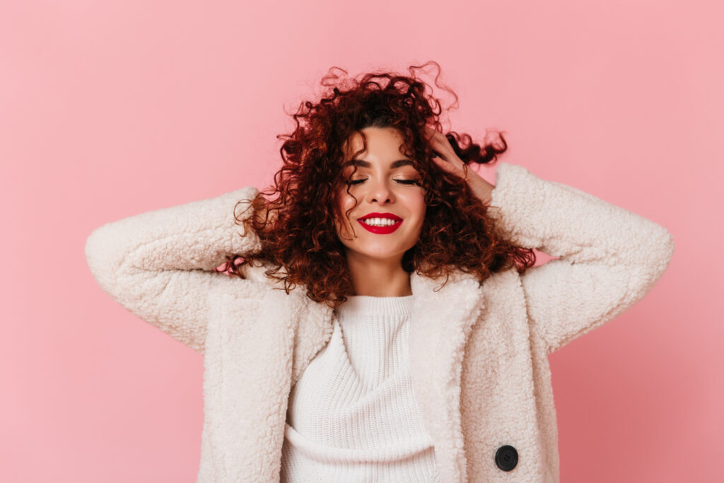 Woman with hands in her curly brown pink hair, standing against a pink backdrop.
