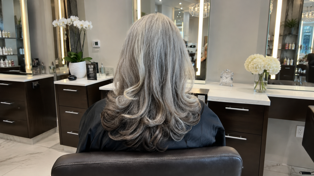 Woman seated in a modern salon chair, seen from behind, with long salt-and-pepper hair styled in a voluminous Hollywood blowout.